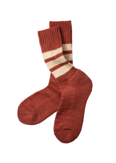 A pair of ribbed knit crew socks in a rust brown color, featuring two horizontal cream stripes near the cuff, displayed on a light gray background.