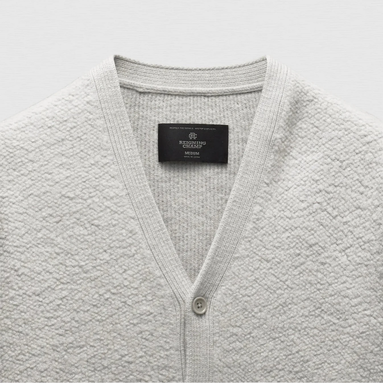 A close-up of a light gray textured knit cardigan sweater with a V-neck and a black neck label reading 'Mikrofibre by Mikael Blom'.