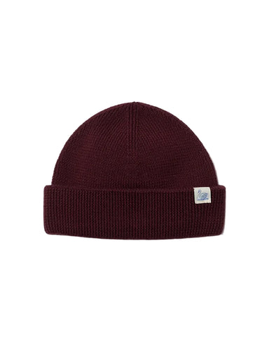 A dark burgundy ribbed knit beanie hat with a small white rectangular brand tag on the side, displayed against a clean off-white background.