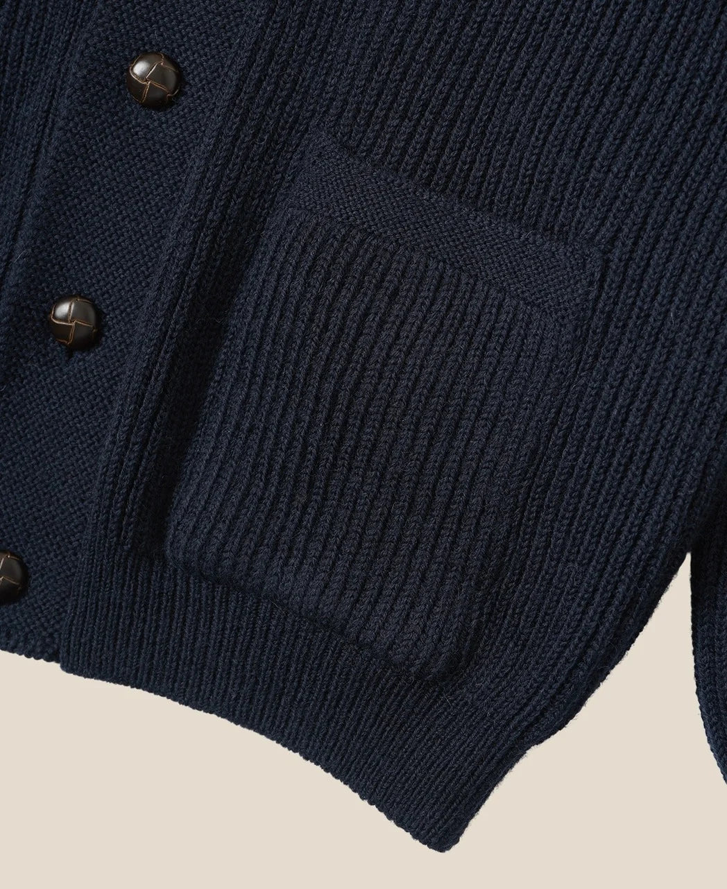 Close-up of a navy blue ribbed knit cardigan sweater showing a front pocket and dark buttons against a light beige background.
